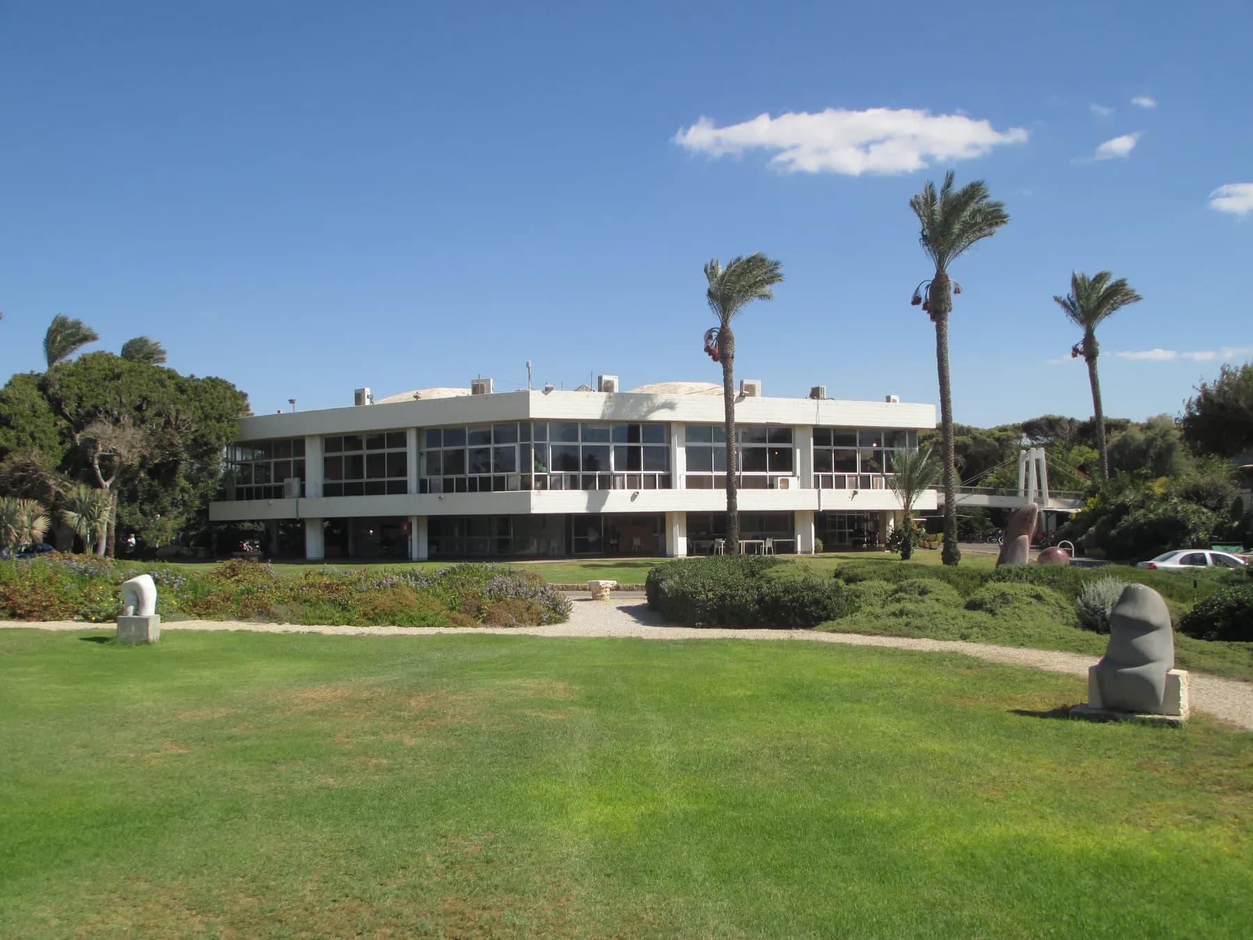 The dining hall of Sdot Yam, a long flat-roofed building with glass walls, behind a wide lawn with palm trees and stone sculptures.