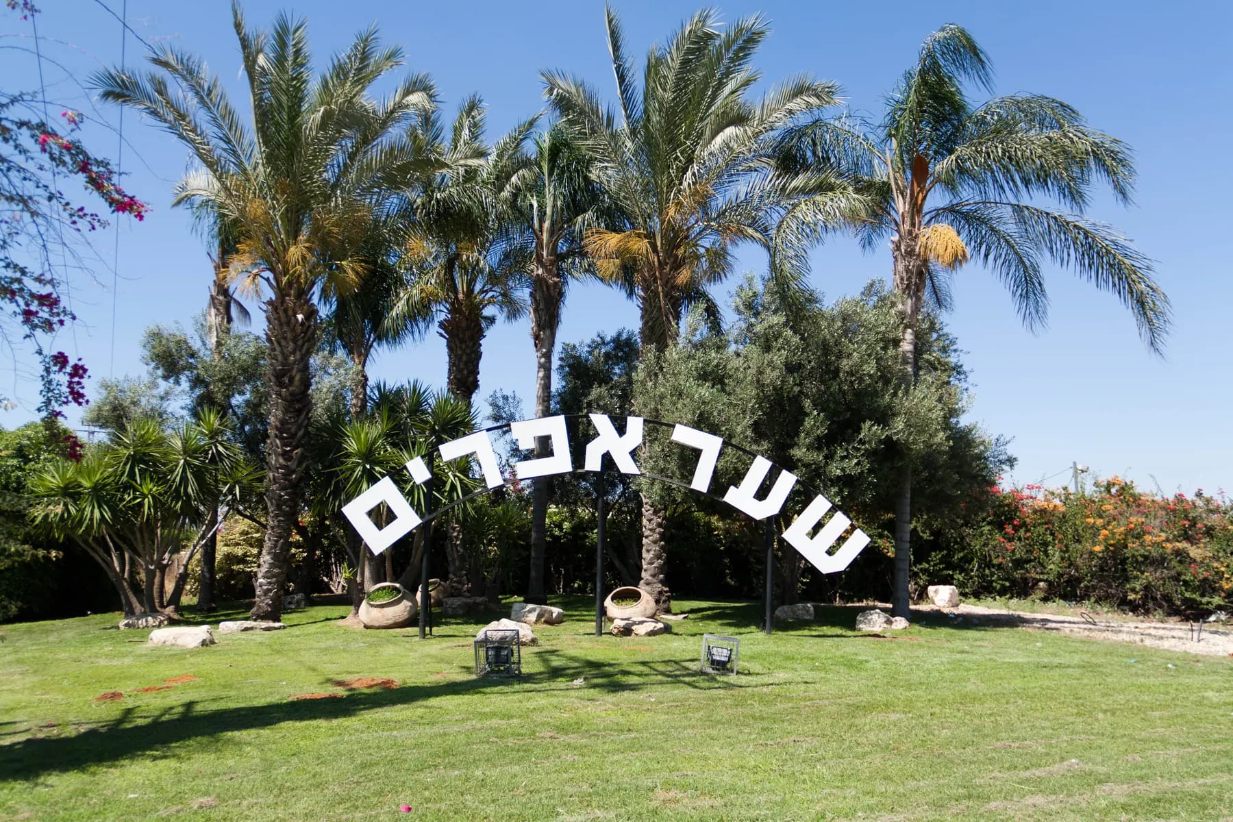 The name Sha'ar Efraim in large white Hebrew letters arched over a lawn at the entrance to the village, with date palms and bushes behind.