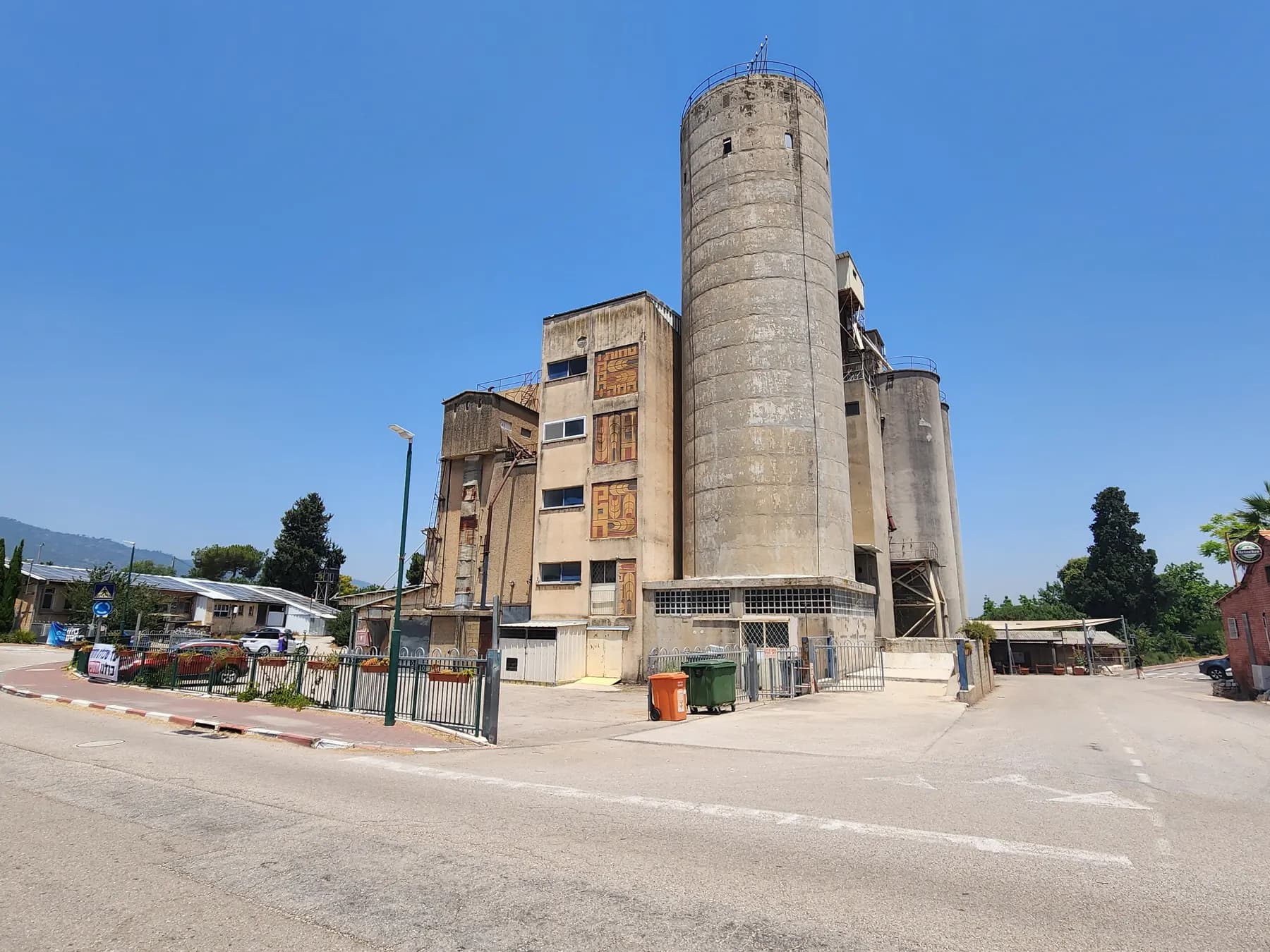 The flour mill of Kibbutz Sha'ar HaAmakim, a tall round concrete silo beside a multi-storey building decorated with ceramic panels, seen from the road.