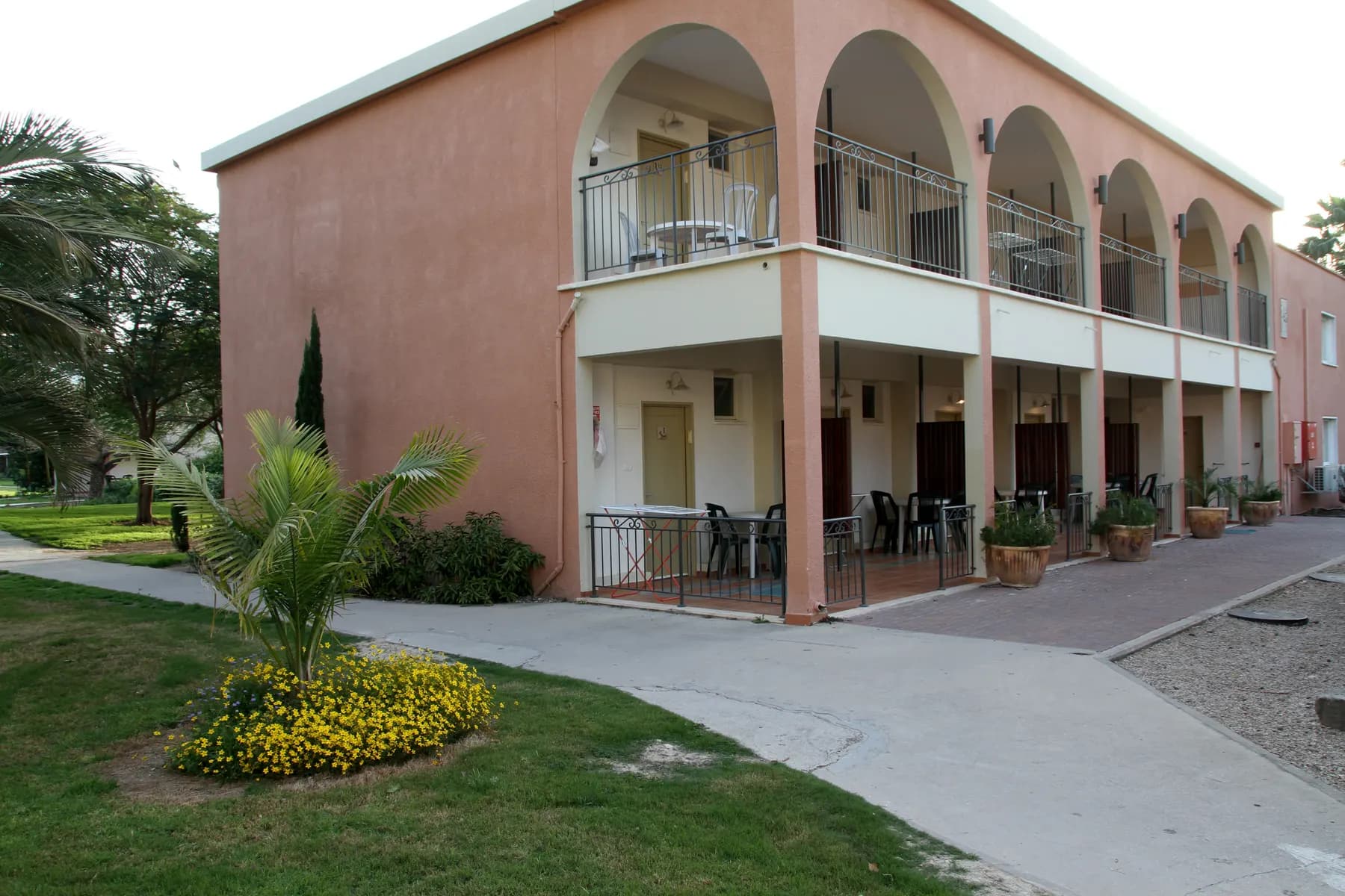 A pink two-storey guest house with arched balconies beside a lawn and a paved path in Sha'ar HaGolan, with palms and flower beds.