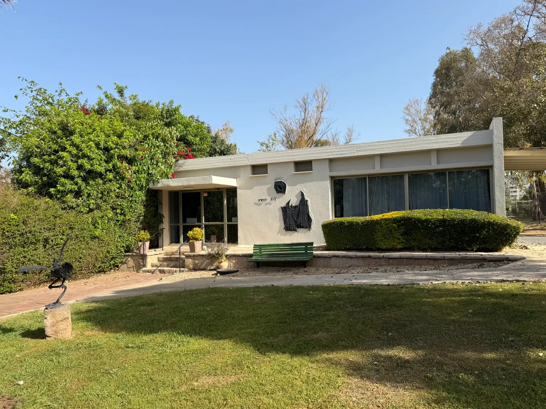 A low white building with a glass front and a dark relief on its wall in Shavei Tzion, behind a lawn with a green bench, trimmed hedges and a small sculpture.