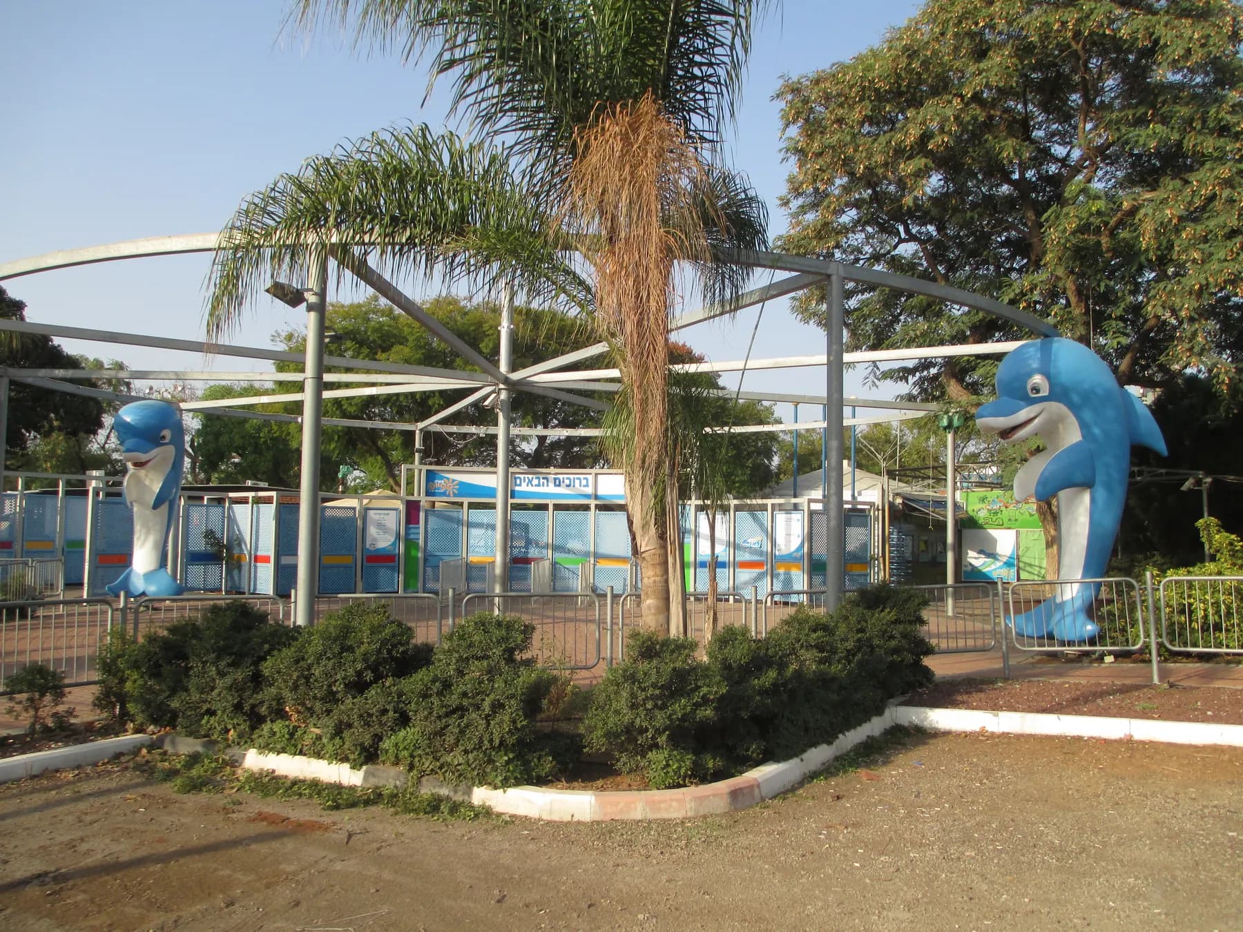 The entrance to the water park in Shefayim, with two blue dolphin statues, a palm tree and a shade canopy.