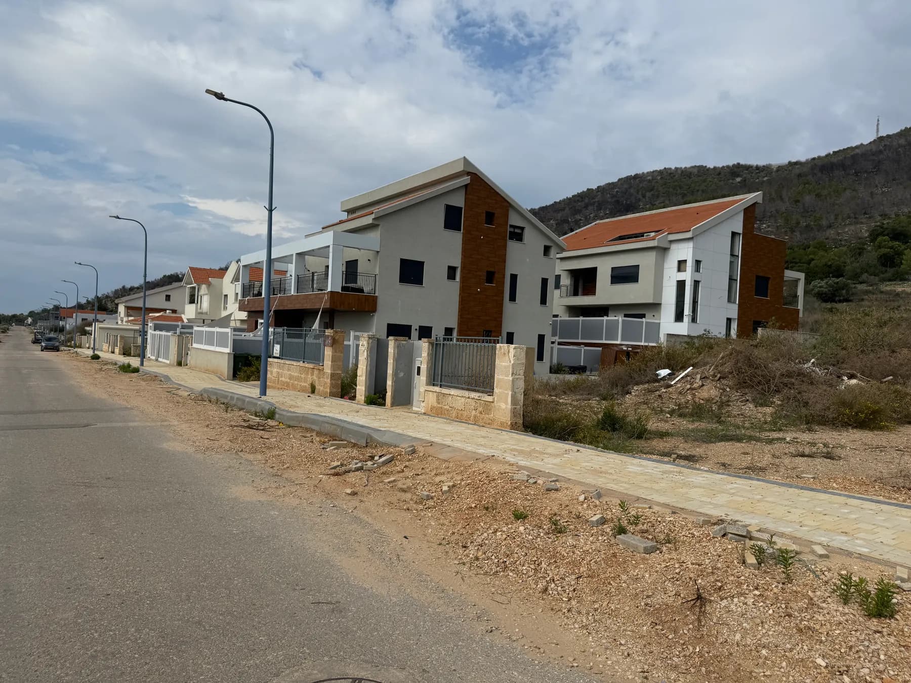 New houses along a quiet street at the foot of a hillside in Shlomi, with street lamps lining the road.