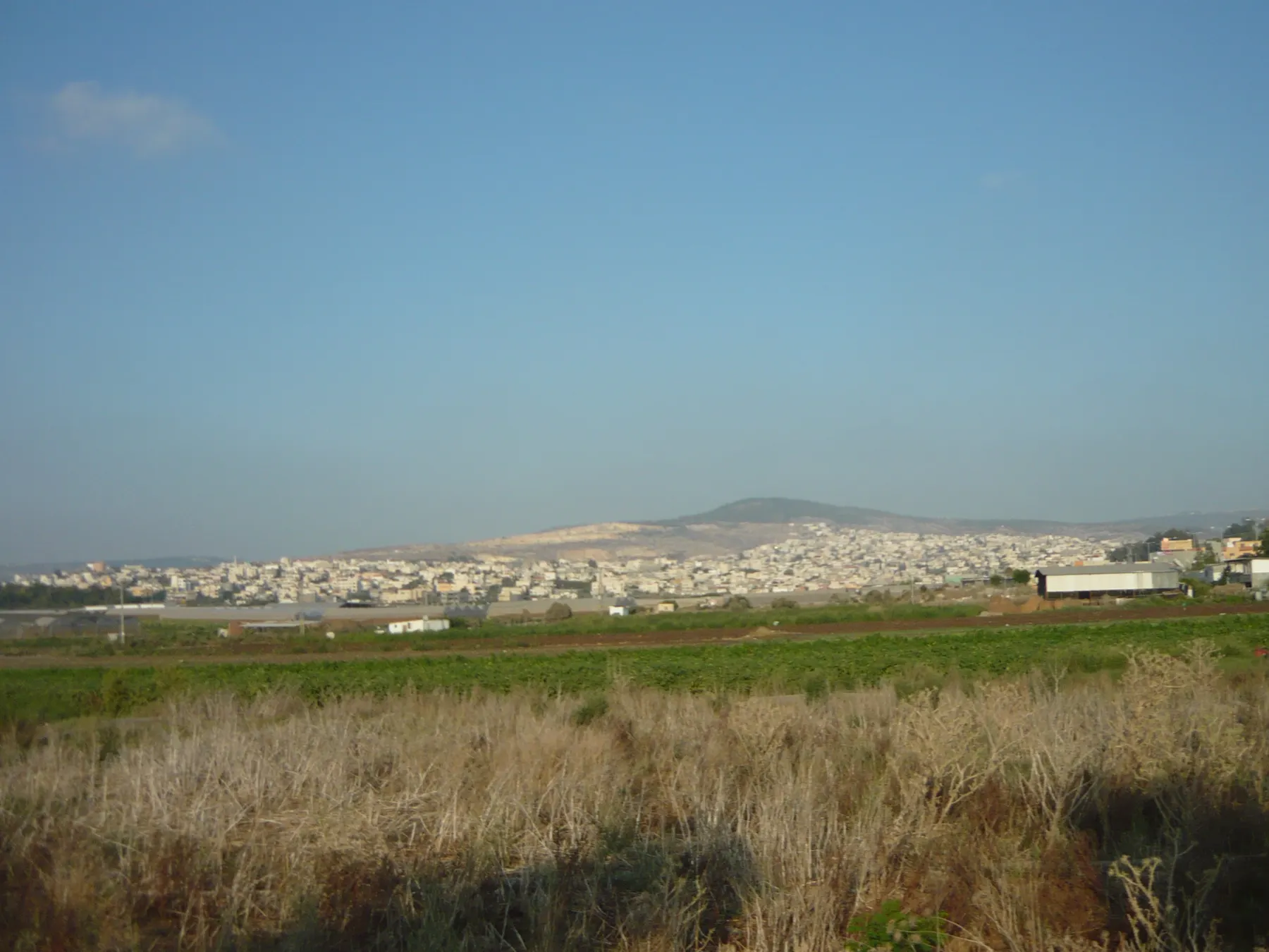 The houses of Tamra spread along the foot of a hill, seen across green fields and dry grass.