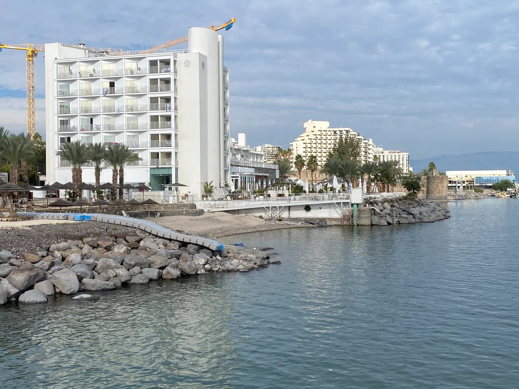 Hotels and palm trees on the shore of the Sea of Galilee in Tiberias, with rocks along the water's edge.