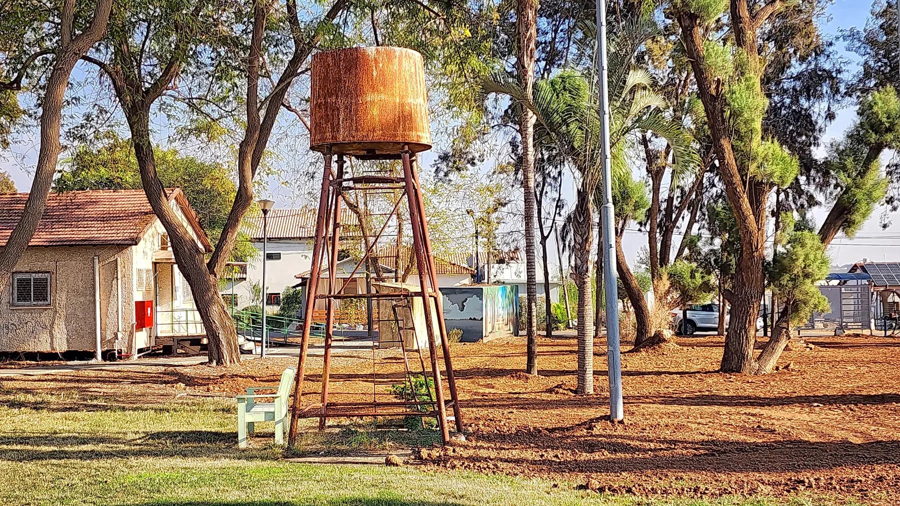 An old rusty water tower on steel legs in a park with trees and a bench in Tkuma, with houses behind.