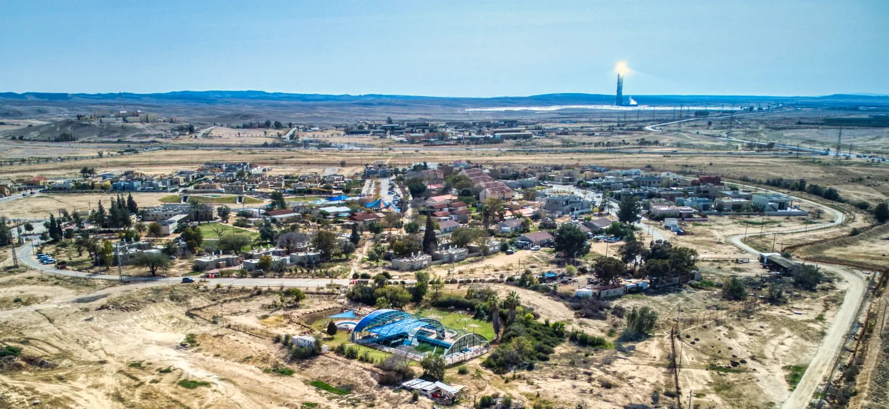 A view from above over the houses and trees of Tlalim in the desert, with a tall tower shining on the horizon.