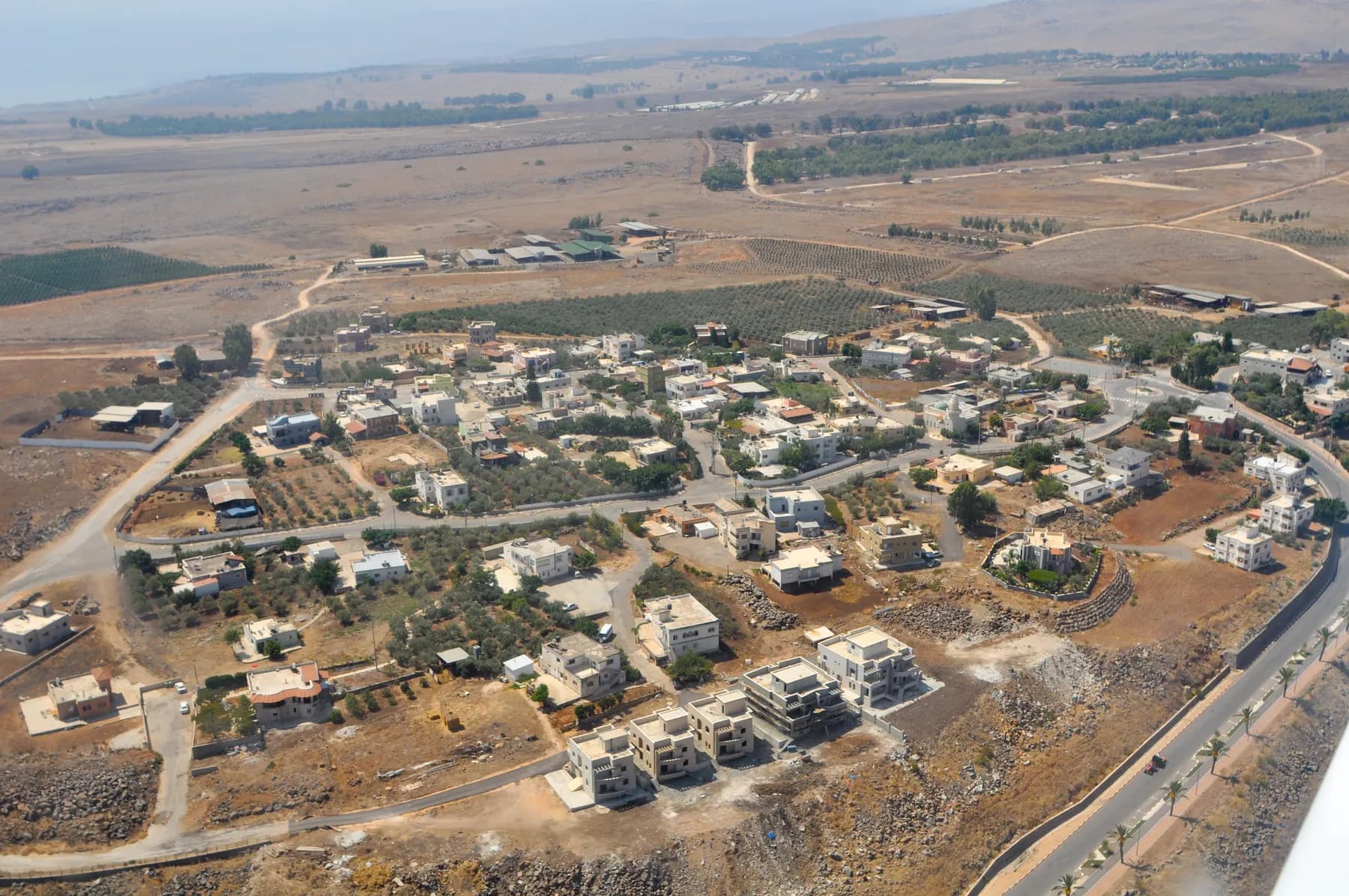 An aerial view of Tuba-Zangariyye, its houses and olive groves spread along winding roads across a dry plateau, with fields and farm buildings beyond.