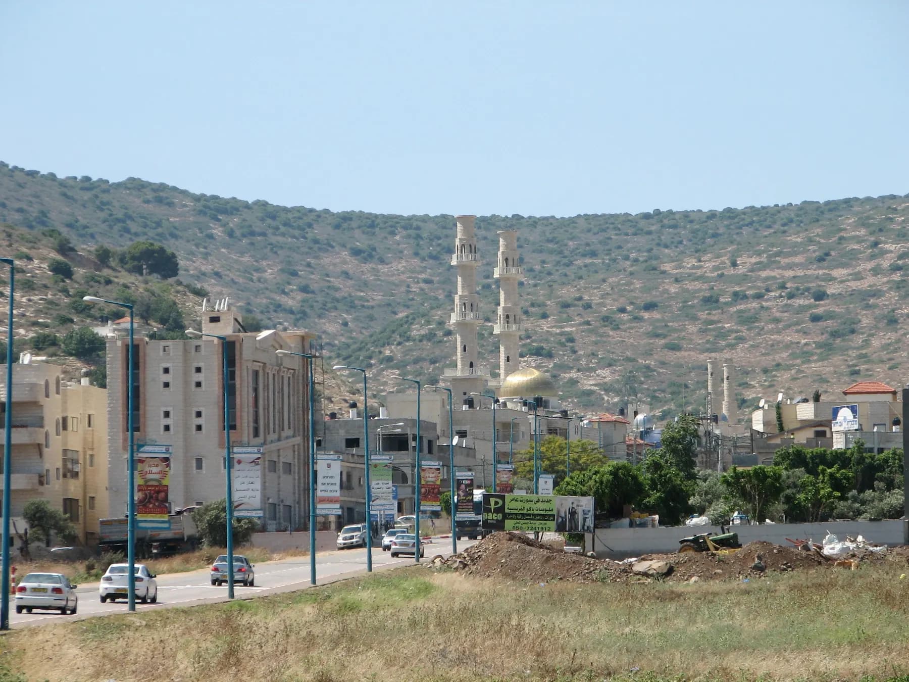 A main road into Tur'an lined with apartment buildings and banners, with a mosque with two tall minarets and a scrubby hillside behind.