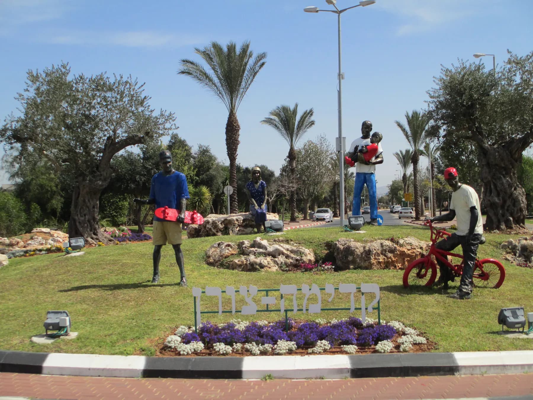 Painted statues of a family and a cyclist on a lawn at the entrance square of Tzoran, with palm and olive trees around.