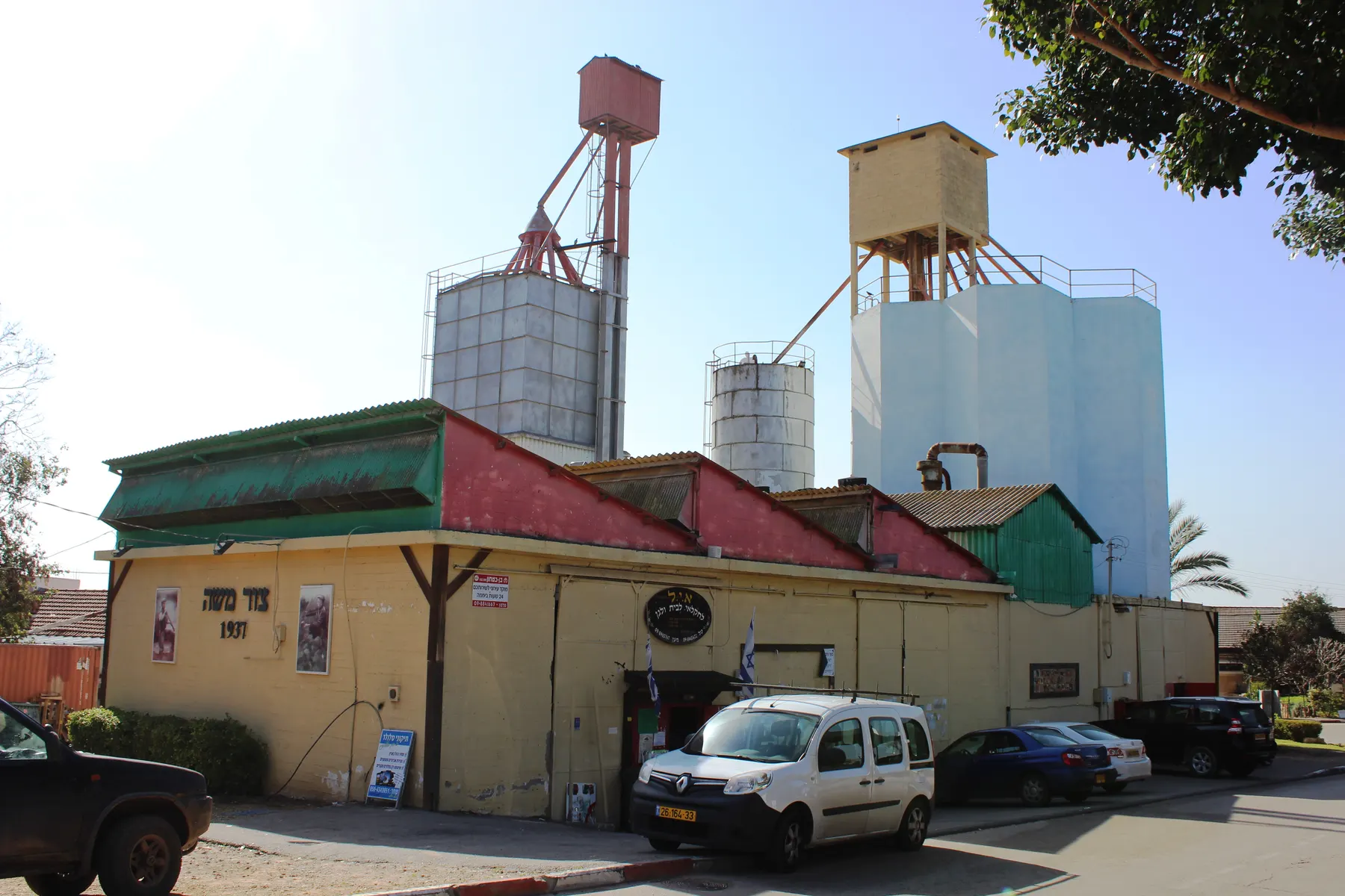 An old yellow farm building marked Tzur Moshe 1937, with tall silos and towers on steel legs behind it and cars parked in front.