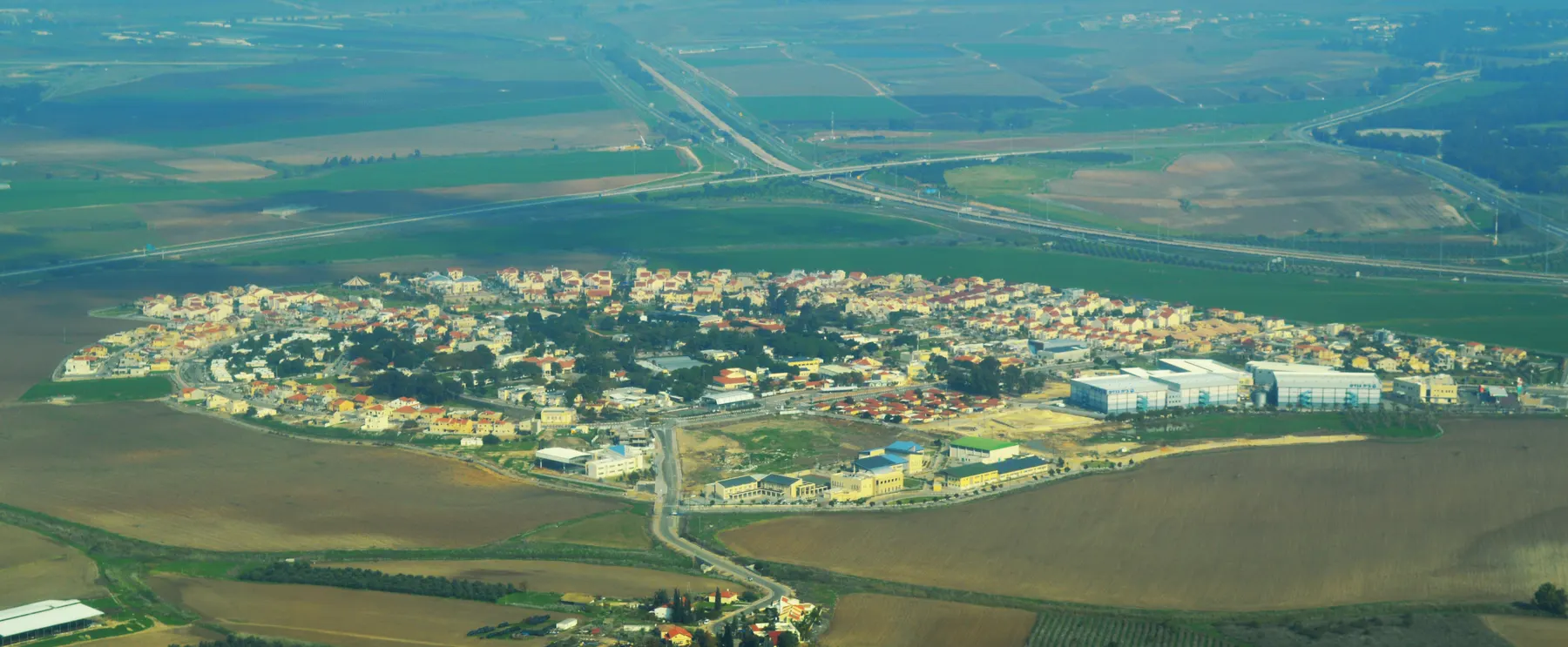 An aerial view of Yad Binyamin, with houses and trees, large industrial buildings on its edge and farmland all around.