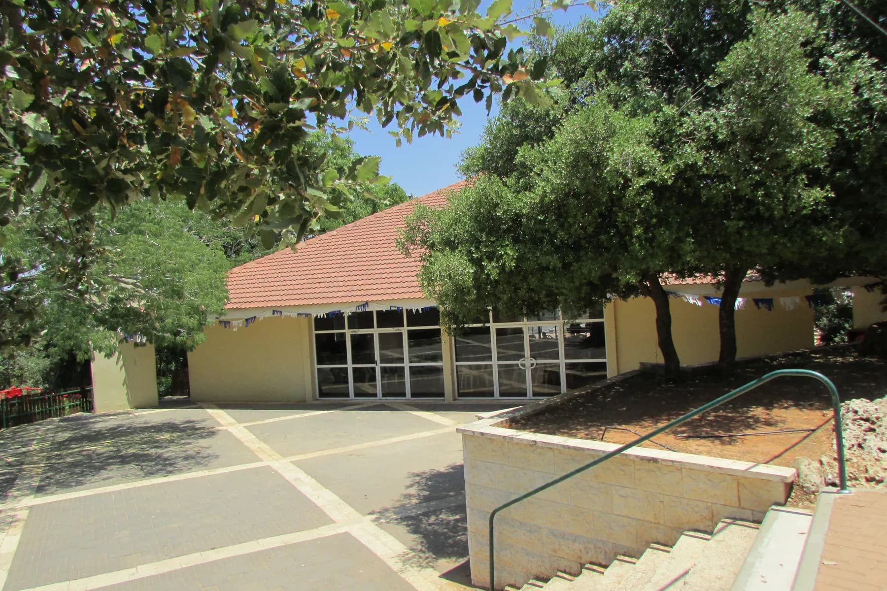A single-storey building with a red tiled roof and glass doors among shady trees in Yakir, with a paved forecourt and steps.