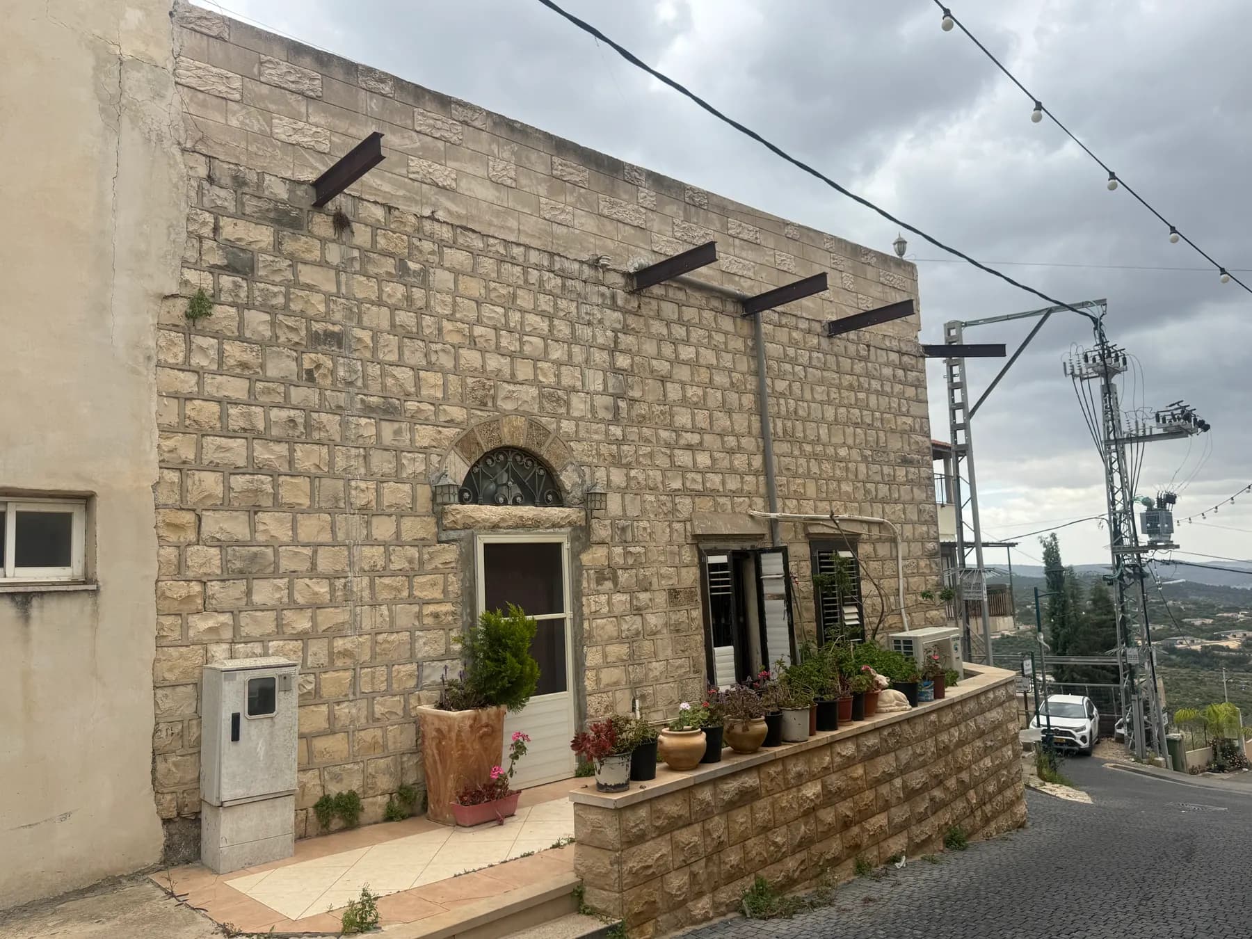 An old stone house with potted plants on a sloping street in the old core of Yarka, with green hills in the distance.