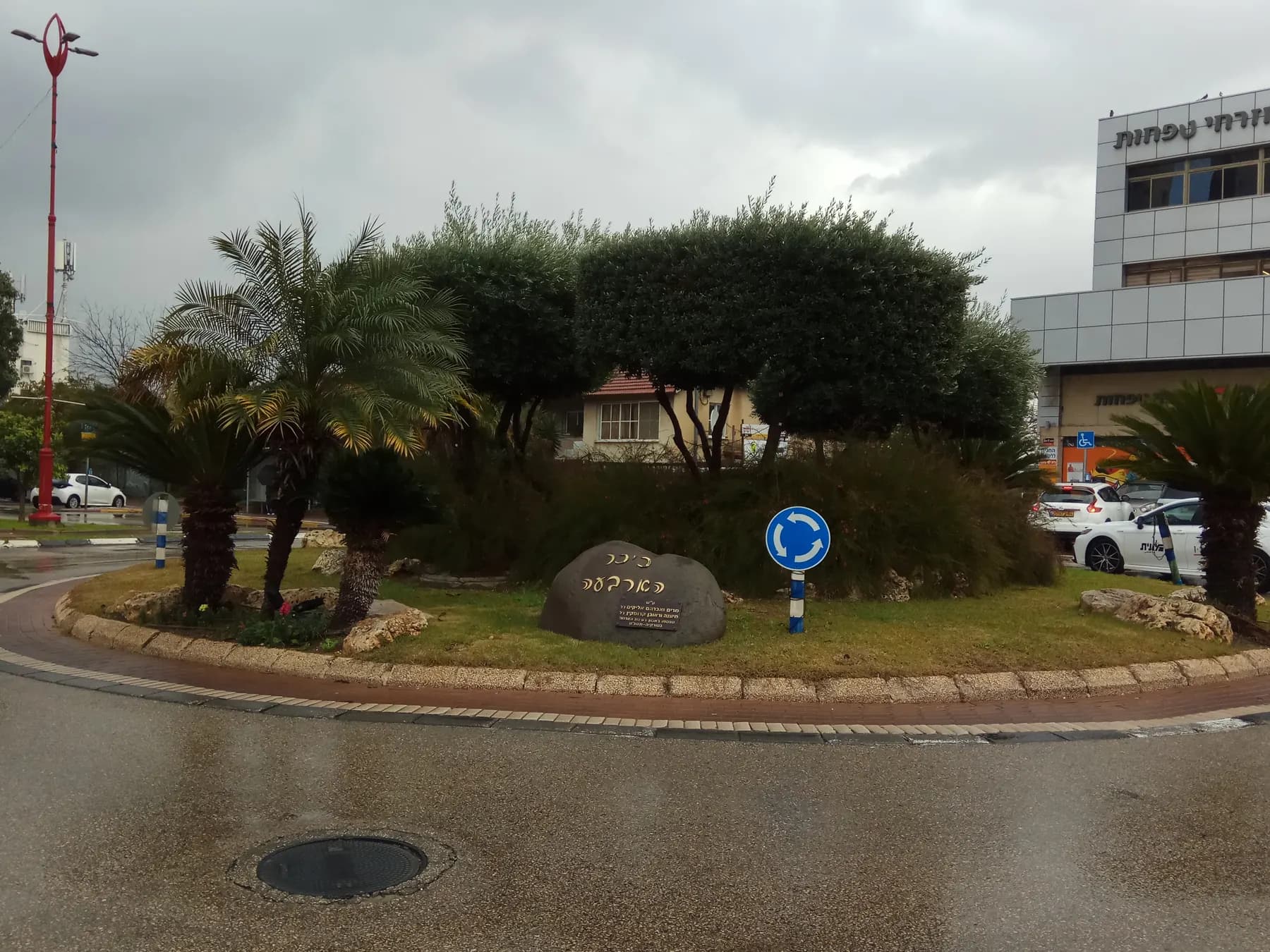 The Four Square roundabout in Yehud on a rainy day, with palms and trees around an engraved stone and an office building behind.