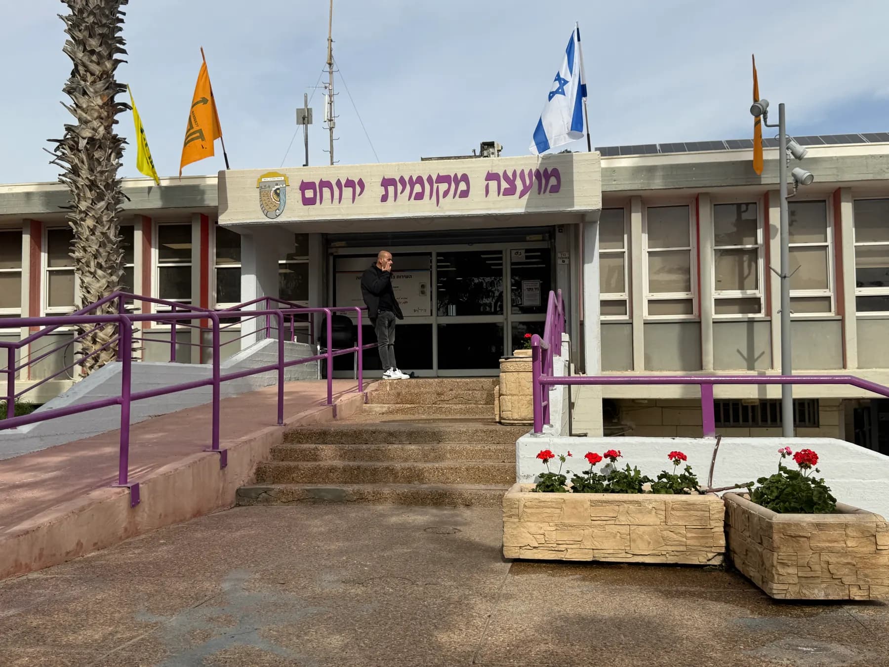The Yeruham local council building, with flags above the entrance, a ramp and planters of red flowers.