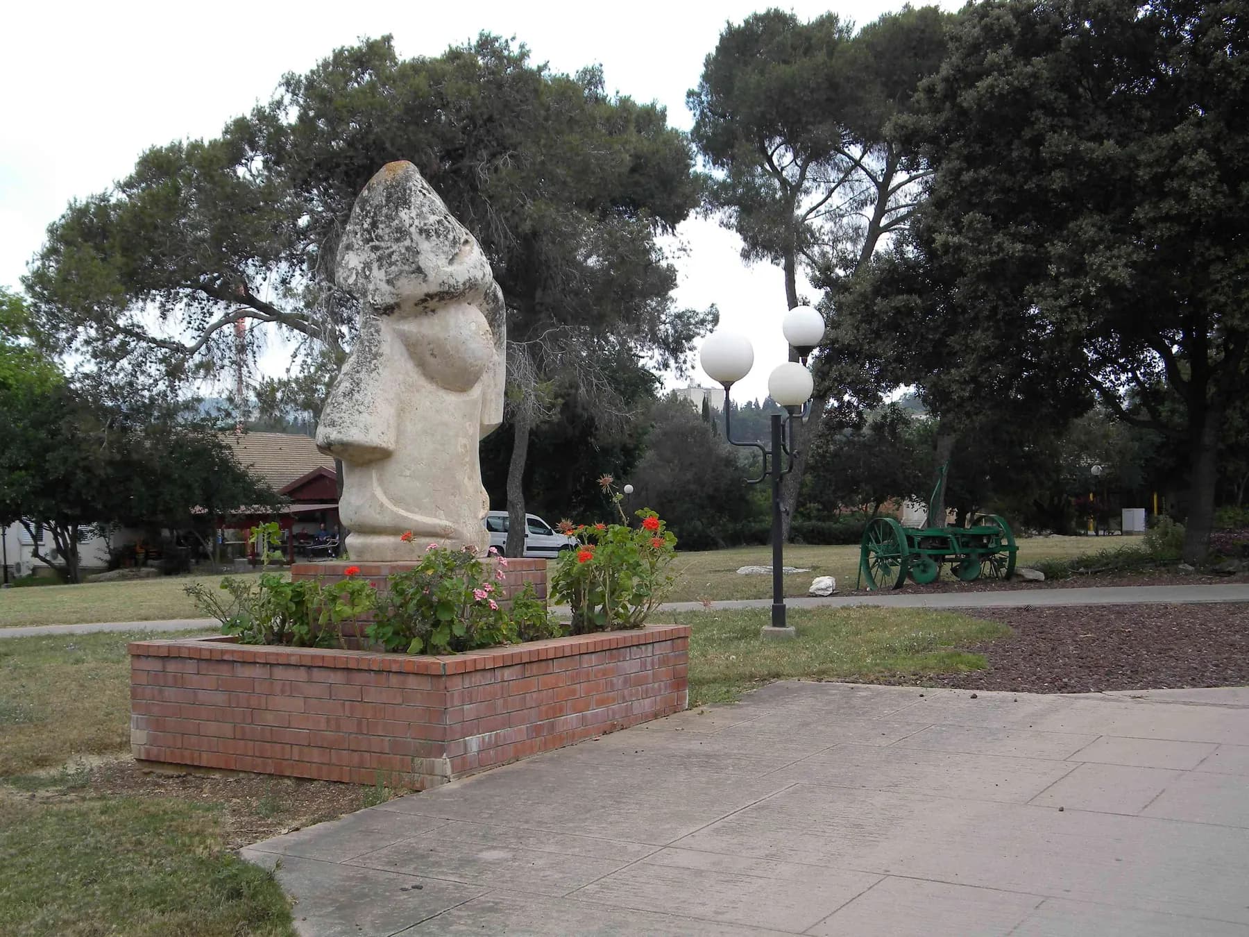 A tall pale stone sculpture on a brick planter of red flowers in Yifat, beside a path with globe street lamps, a lawn and shade trees, with an old green farm machine on wheels behind.