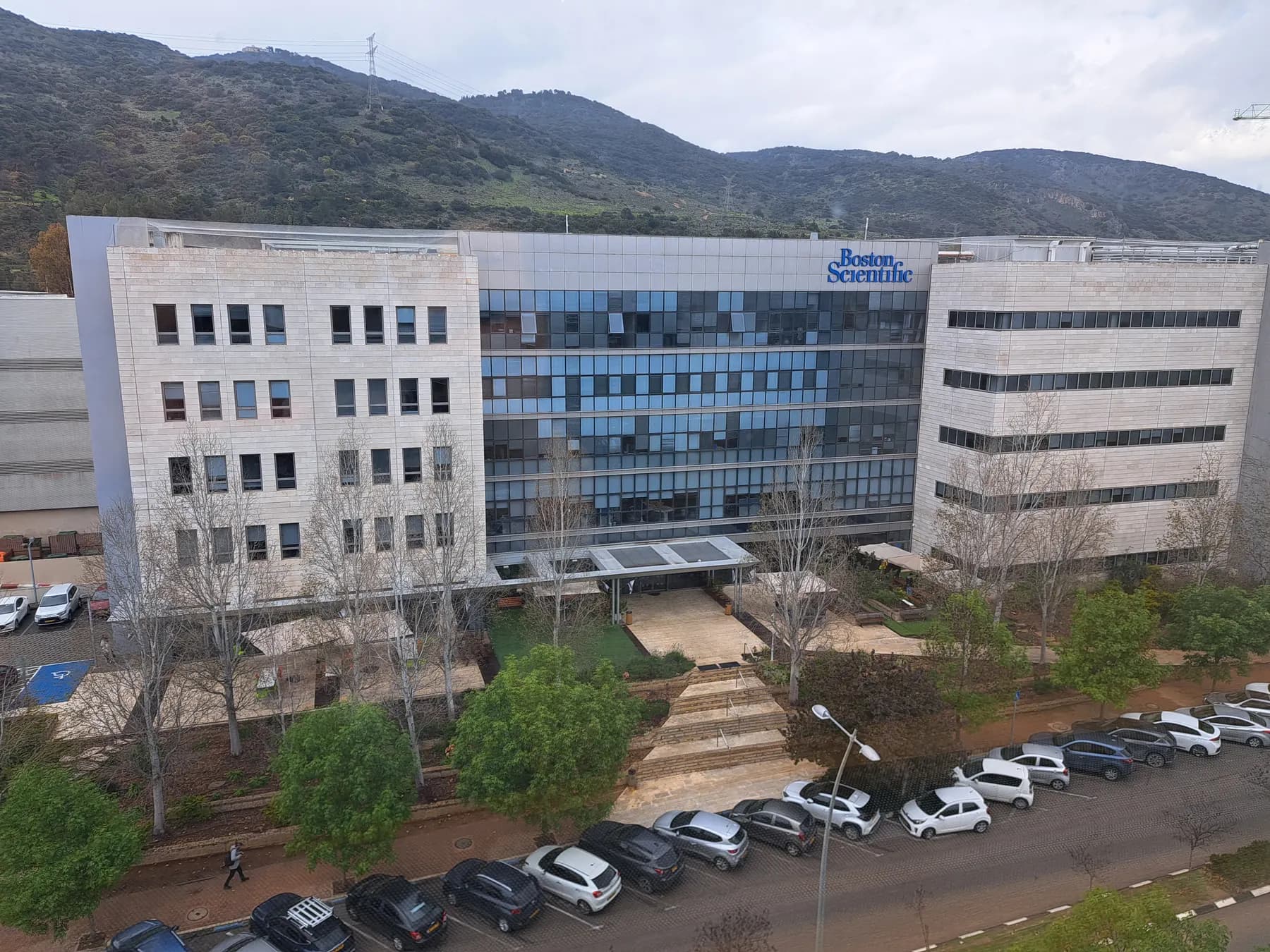A glass and stone office building in Yokneam with cars parked in front and wooded hills behind.