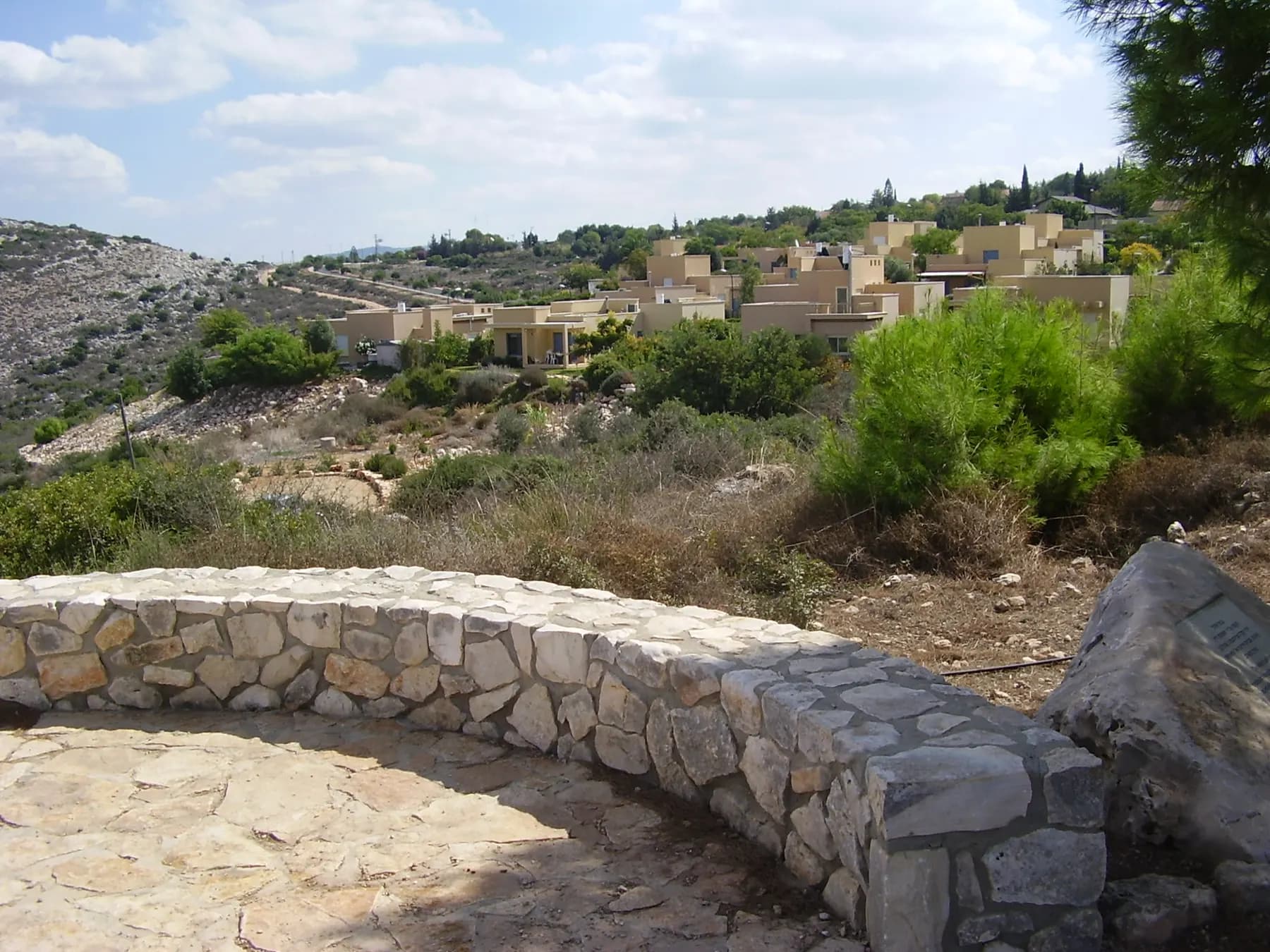 Houses with pale walls on a hillside in Yuvalim, seen from a lookout with a low stone wall, with green bushes and rocky slopes around.