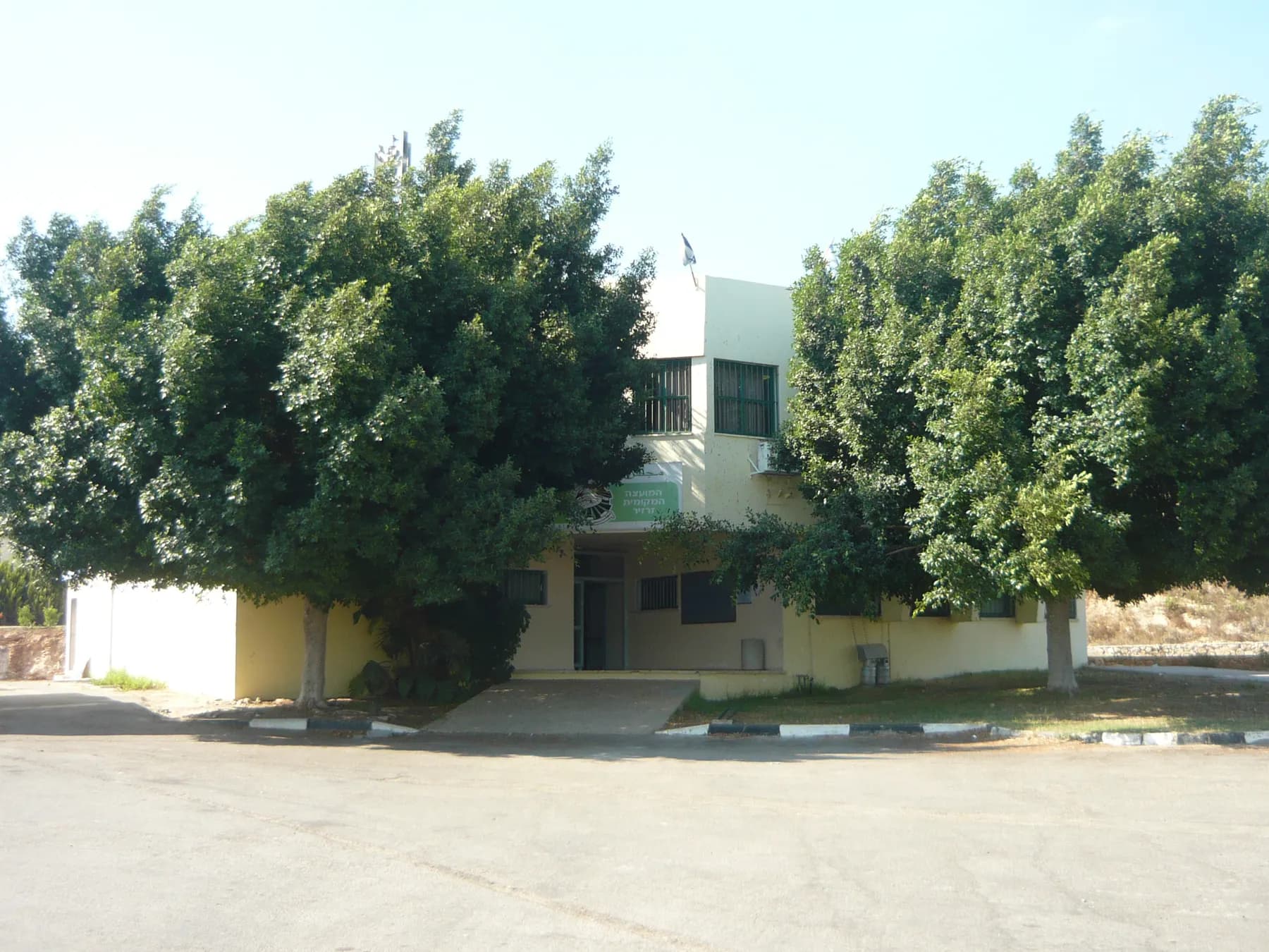 The local council building in Zarzir, half hidden behind two large leafy trees, with an empty paved forecourt in front.