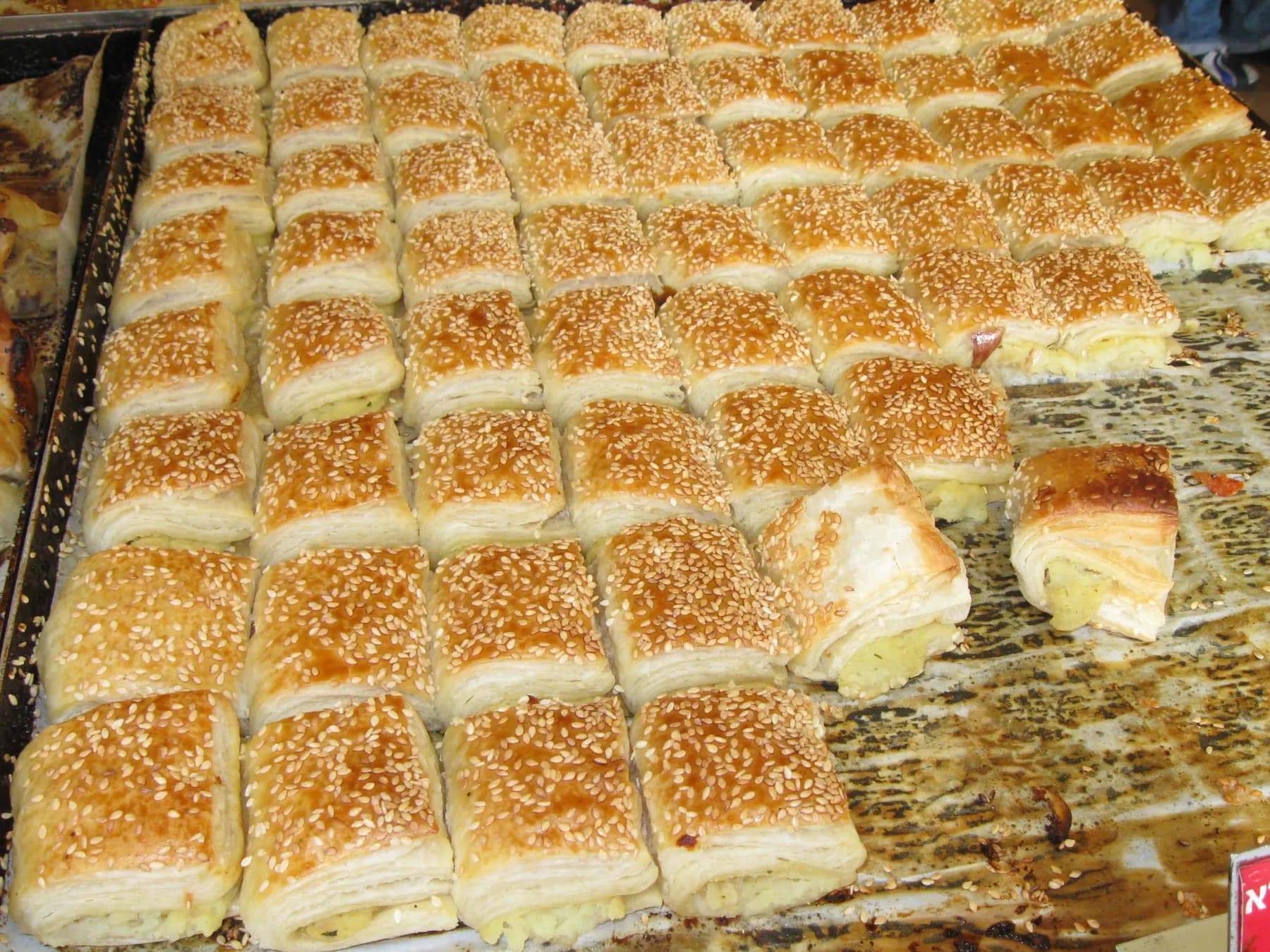 Rows of square sesame bourekas on a baking tray, one cut open to show the potato filling.