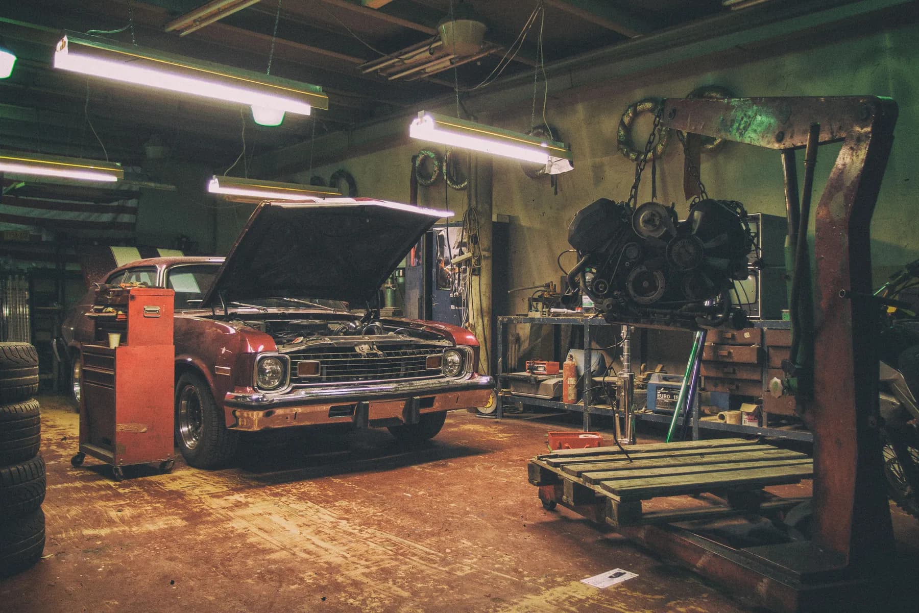 A car with its bonnet open in a working garage, beside an engine on a hoist and stacks of tyres