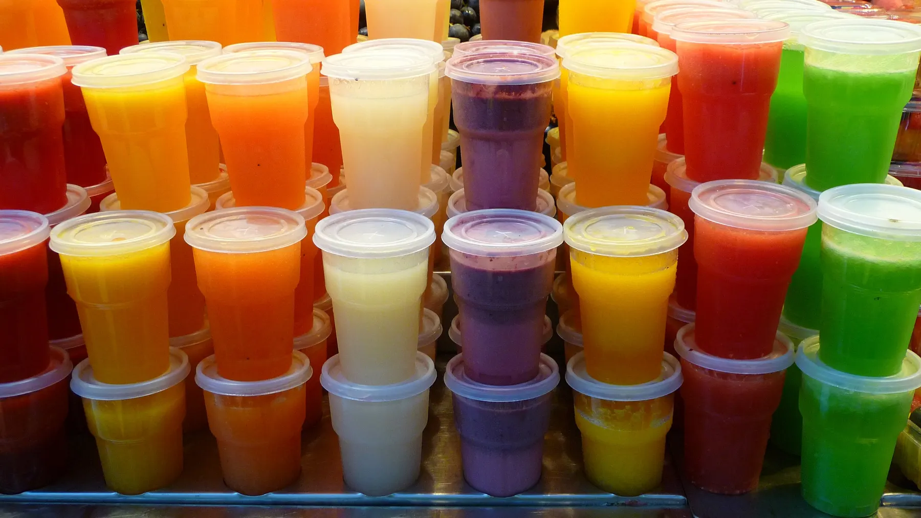 Rows of fruit smoothies in lidded cups, orange, red, green and purple, on a market counter