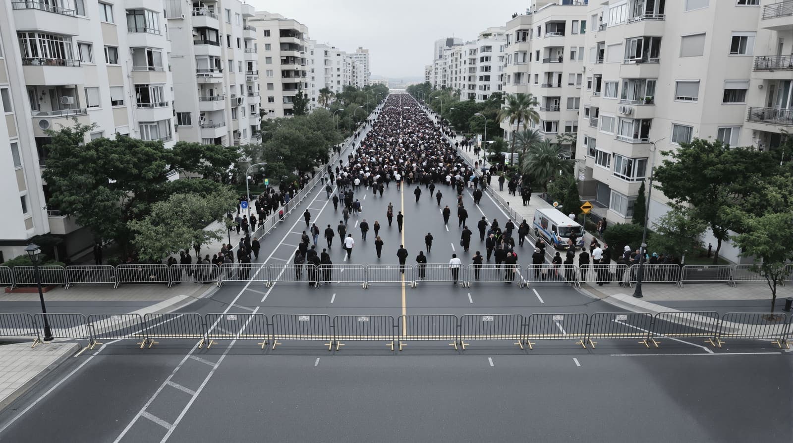 Illustration of a closed Netanya street seen from above, steel barriers holding back a vast crowd of mourners filling the road between apartment blocks.