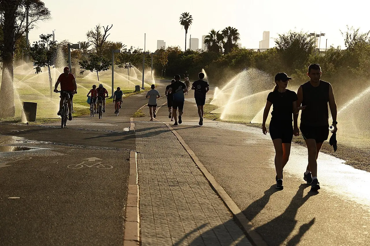 Walkers and cyclists on a path in HaYarkon Park on a Saturday morning, sprinklers running on the lawns